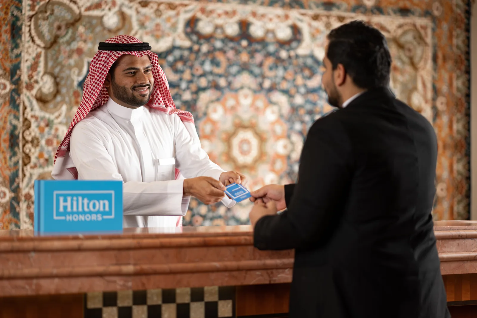 Hotel front-desk welcome — Saudi host handing a Hilton Honors key card to a guest